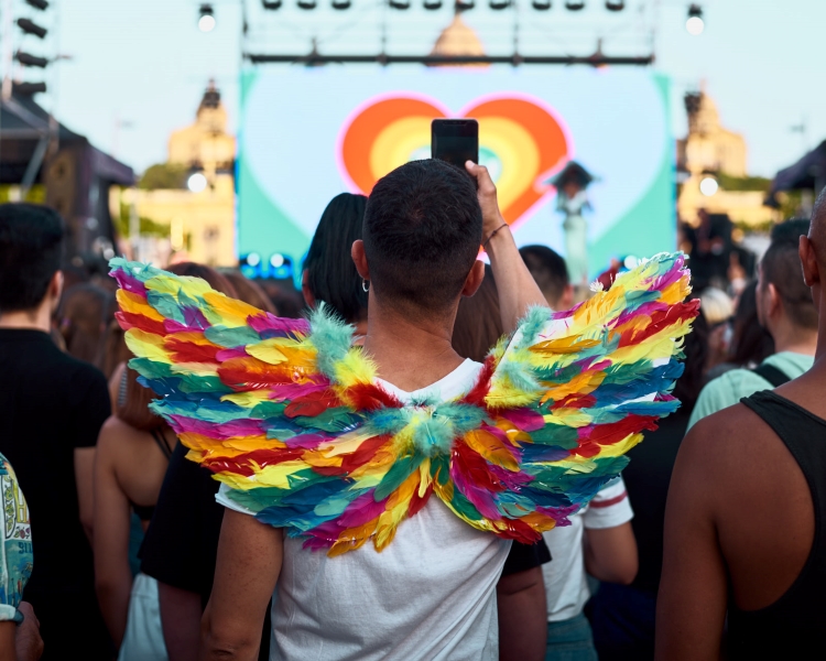 A man holding his phone wearing colorful wings during a Pride! Barcelona event on June 28, 2019 (by Pride! Barcelona)